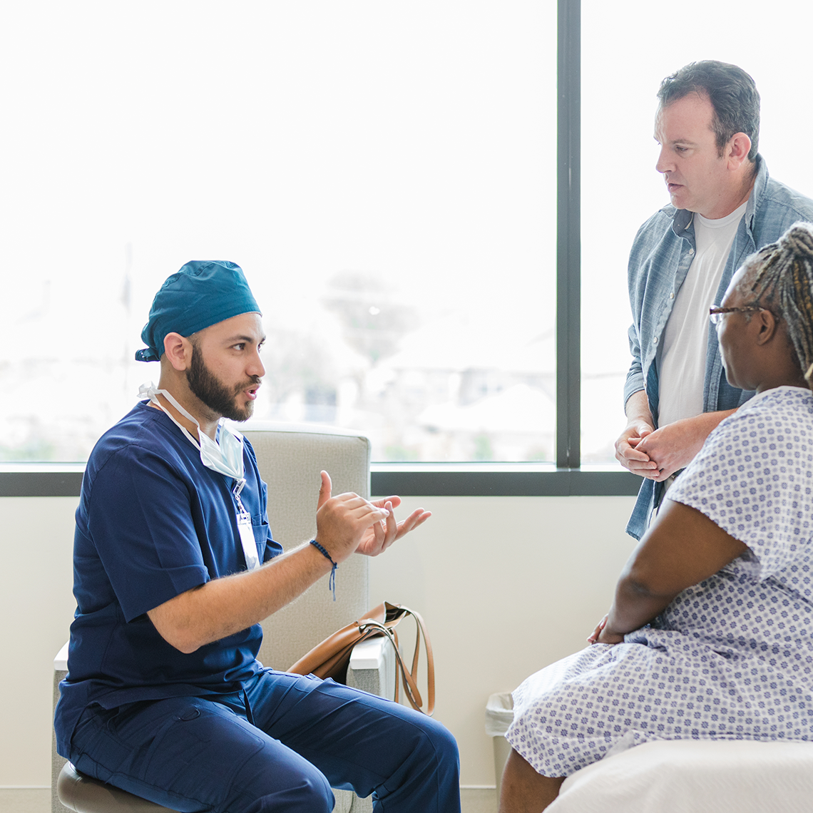 person in scrubs talks to a woman in a johnny who is seated on a hospital bed; a man stands beside her