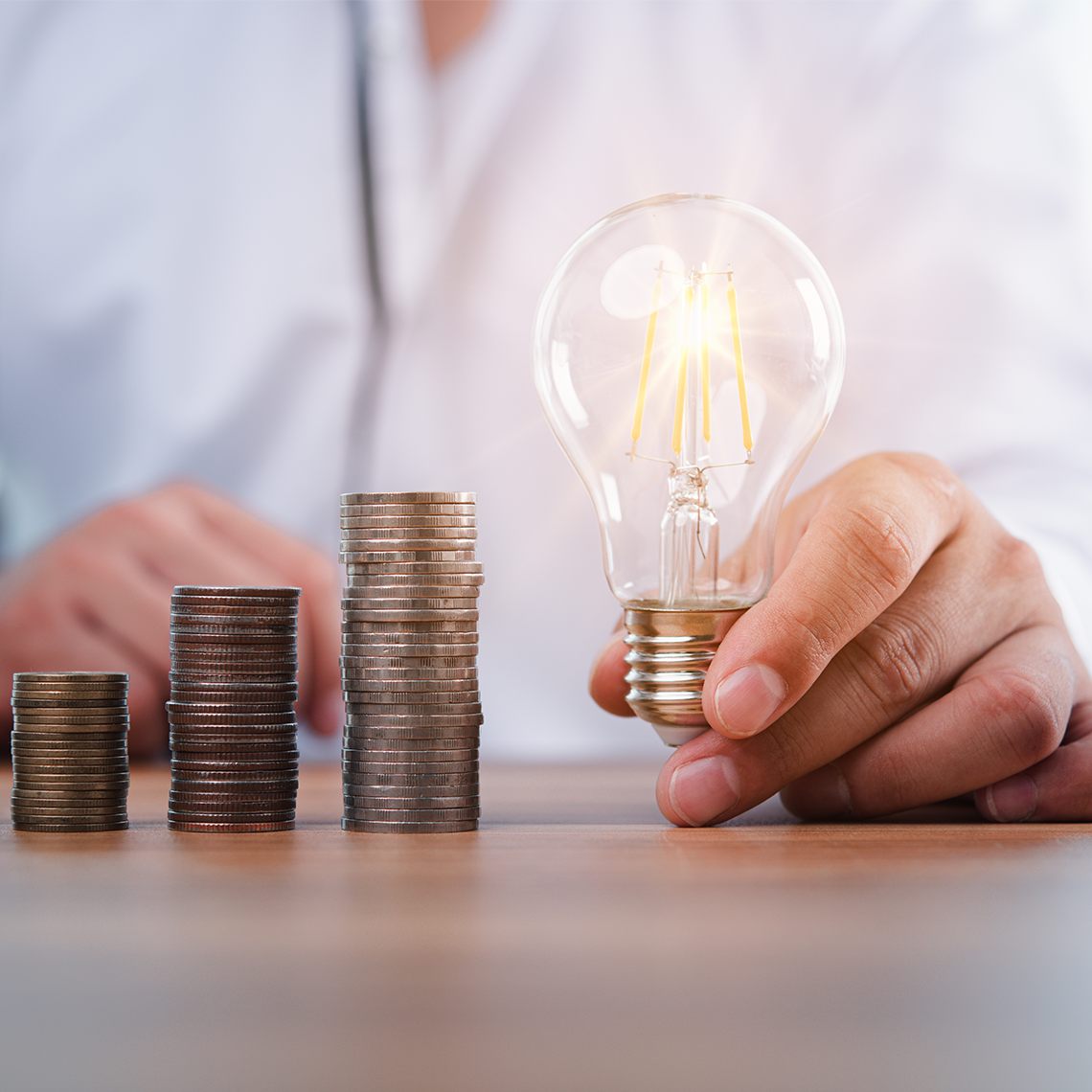 Close up of four increasing stacks of coins on a desk in front of a person wearing white. They are holding a lightbulb to the right of the tallest coin stack.